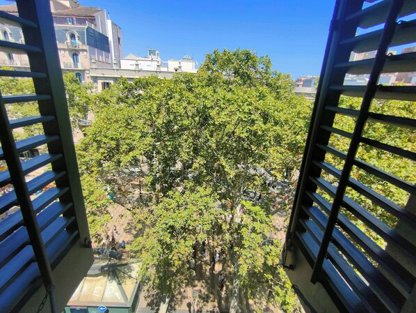 A view from a balcony with metal railings, a tree in the foreground, and buildings in the background.