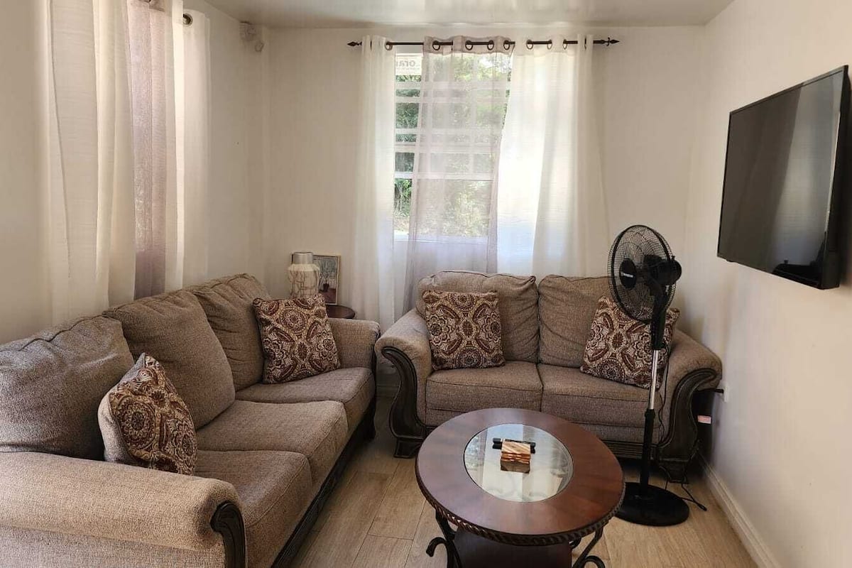 A living room with two sofas, a coffee table, a standing fan, and a flat-screen TV mounted on the wall.