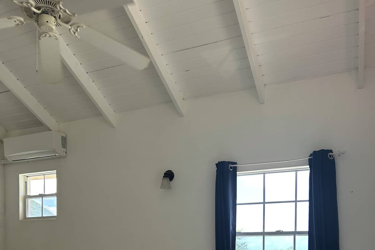 A hotel room with a bed, a window with curtains, a ceiling fan, and a small window with a view.