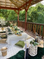 Uma área de refeições ao ar livre com um deck de madeira coberto, toalhas de mesa brancas e guardanapos verdes.