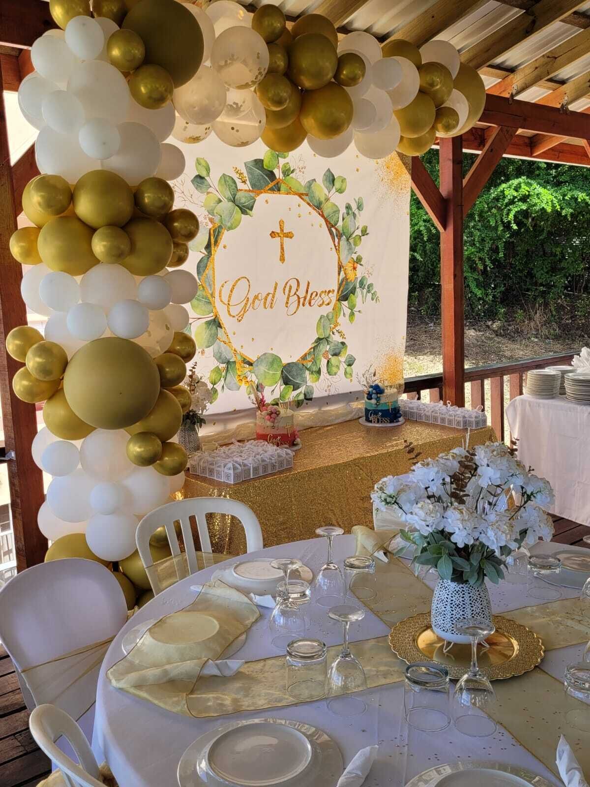 A decorated outdoor dining area with a white and gold color scheme, featuring a table set for a meal, a floral arrangement, and a backdrop with a cross and the phrase "God Bless."