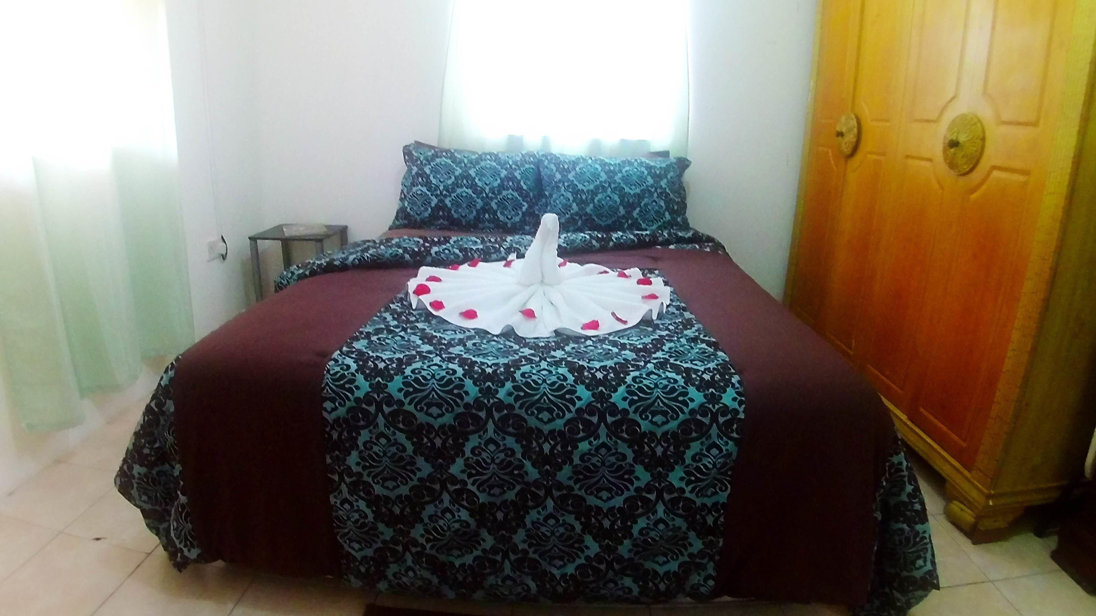 Una cama bien hecha con una colcha estampada y almohadas, una mesita de noche pequeña y un armario de madera.