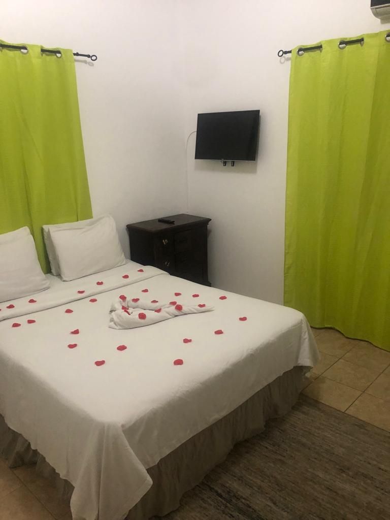 A hotel room with a bed, green curtains, a TV mounted on the wall, and a small nightstand.
