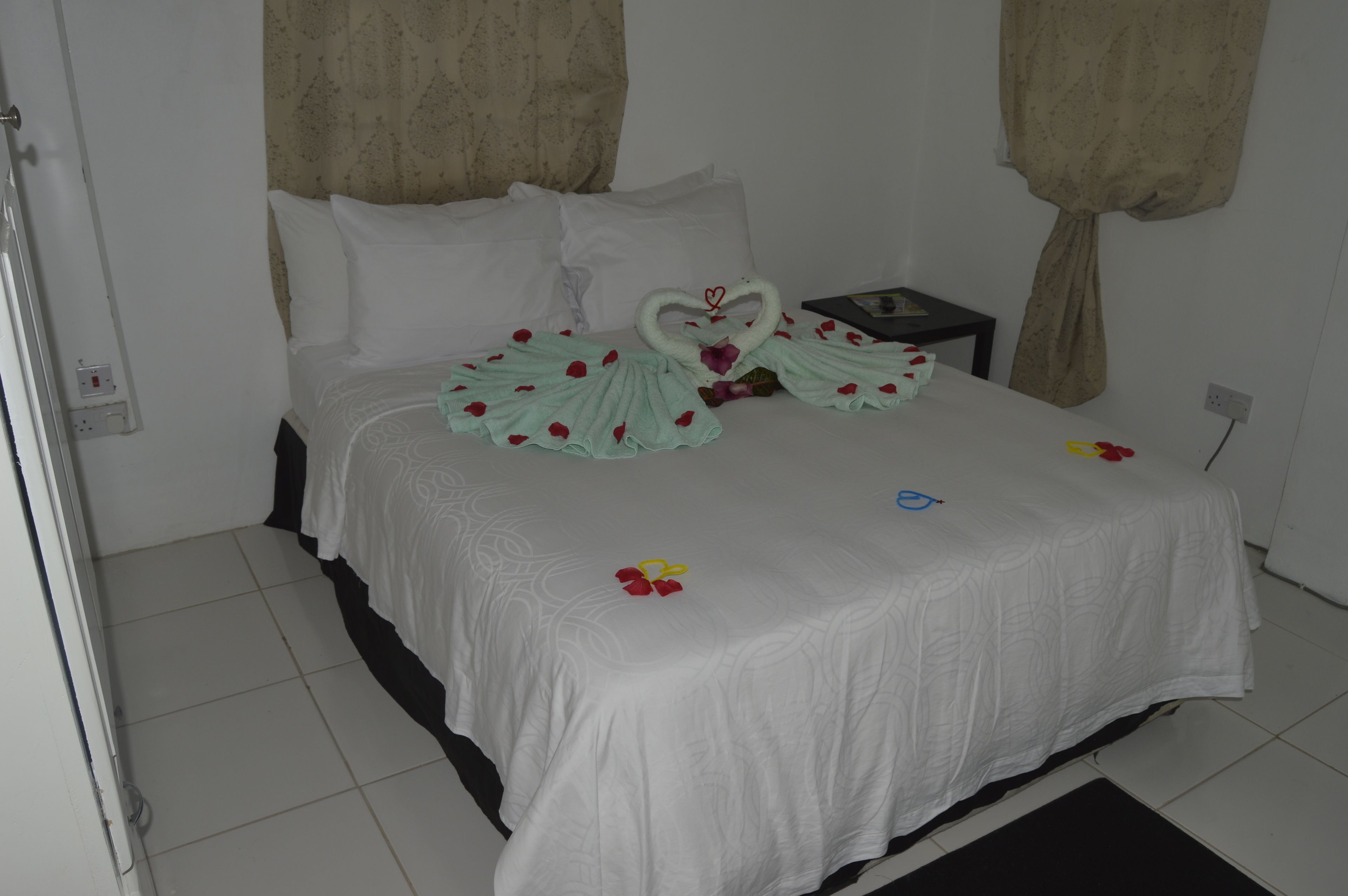 Cama bem arrumada com colcha branca, dois cisnes de tecido e pétalas de rosa espalhadas.