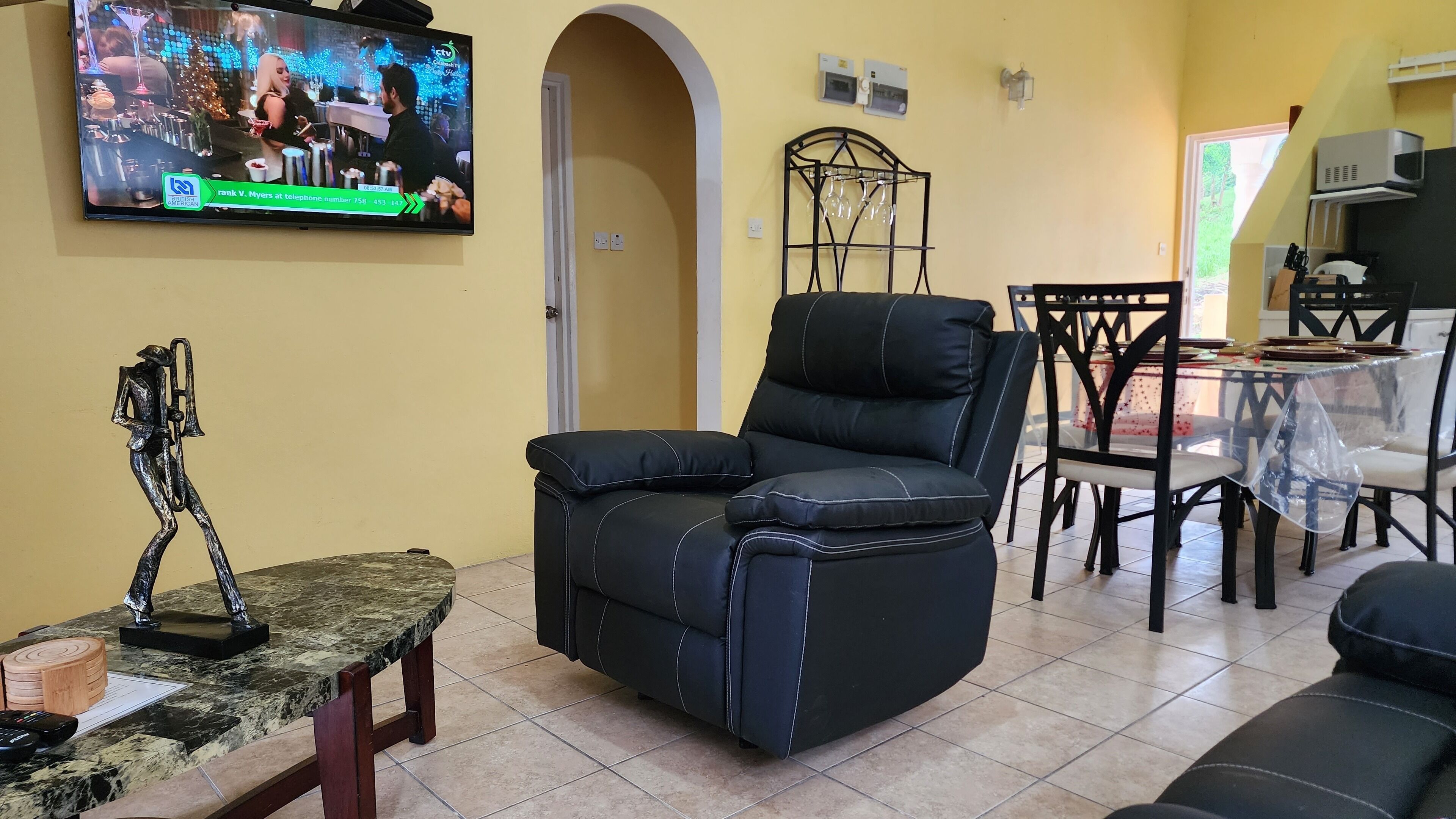A living room with a black leather recliner, a marble coffee table, a TV mounted on the wall, a dining table with chairs, and a decorative metal sculpture.