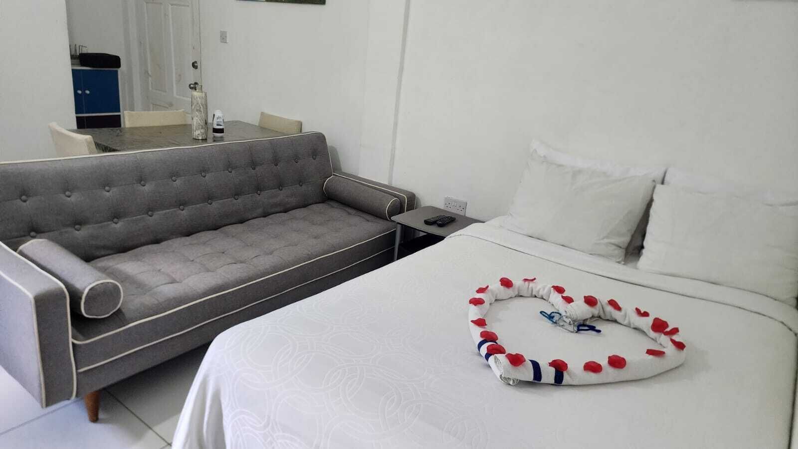 A hotel room with a grey sofa, a neatly made bed with a heart-shaped pillow, and a bedside table with a remote control.