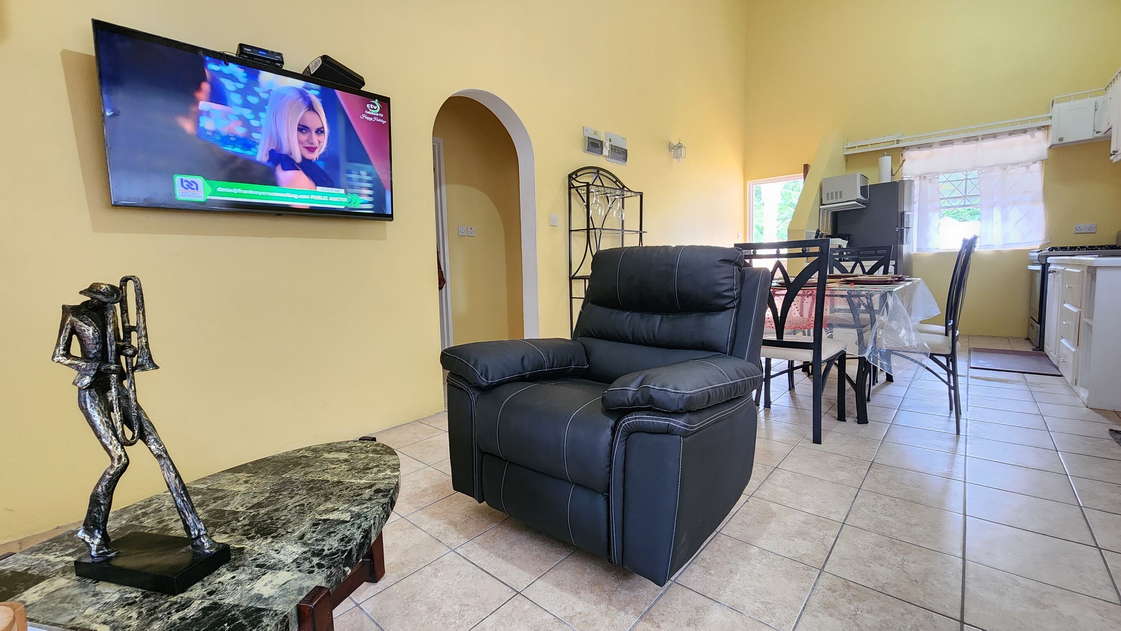 A living room with a black leather sofa, a marble table, a television, and a kitchen area with a stove and cabinets.