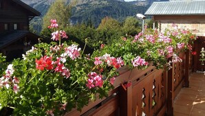 Uma varanda de madeira adornada com flores rosa e vermelhas, com vista para uma paisagem montanhosa.