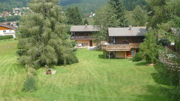A residential area with two houses, one with a balcony and another with a deck, surrounded by lush greenery and mountains in the background.