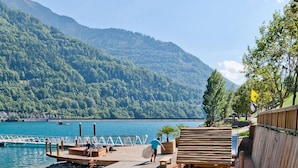 A wooden deck with benches overlooking a lake, a wooden fence, and a dock with boats. - ALLEMONT Chalet des Galline DOMAINE VAUJANY ALPE D HUEZ  (Allemond)