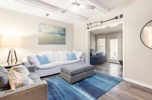 Een moderne woonkamer met een grijze bank, voetenbankje en een blauw tapijt. Aan de muur hangt een schilderij en er is een plafondventilator.