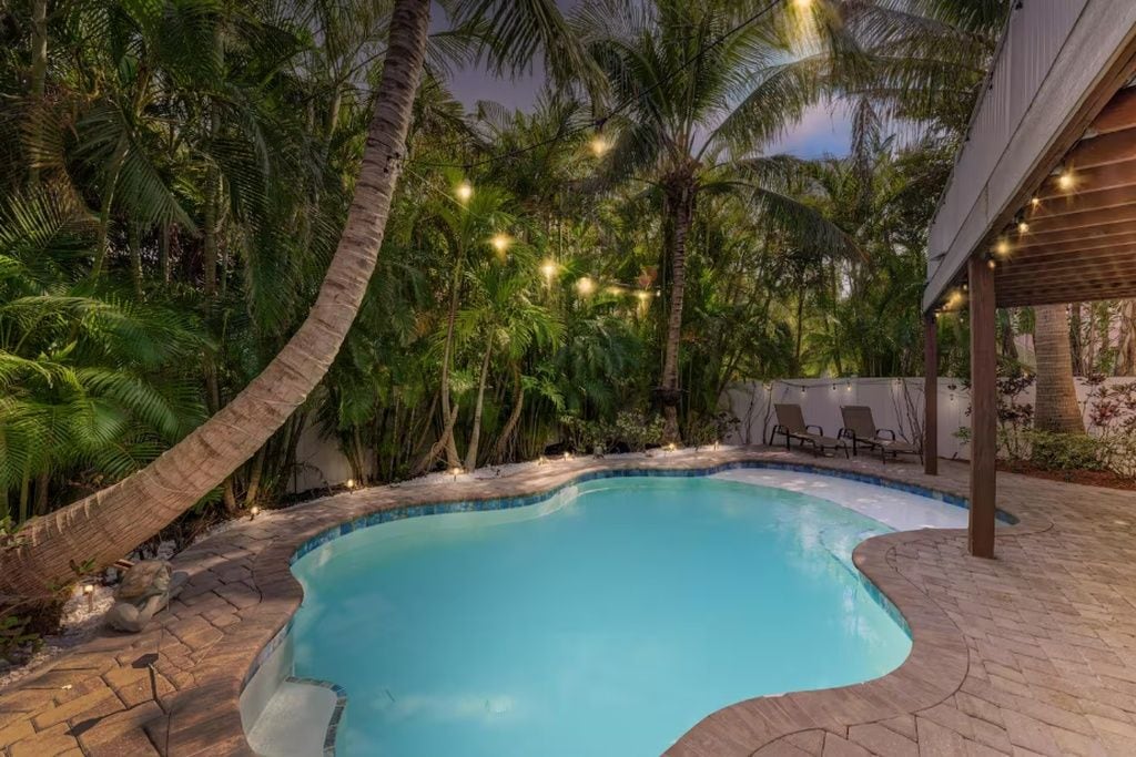 A swimming pool surrounded by palm trees and a covered seating area with string lights.