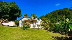 A house with a white exterior and a green roof, surrounded by lush greenery and a clear blue sky. - Bungalows with swimming pool with breathtaking sea and mountain views for 4 people each (BOUILLANTE)