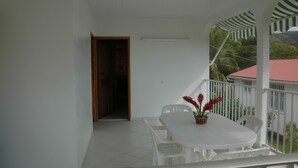 A balcony with a white table and chairs, a wooden door, and a view of a red-roofed building and greenery. - Bungalows with swimming pool with breathtaking sea and mountain views for 4 people each (BOUILLANTE)