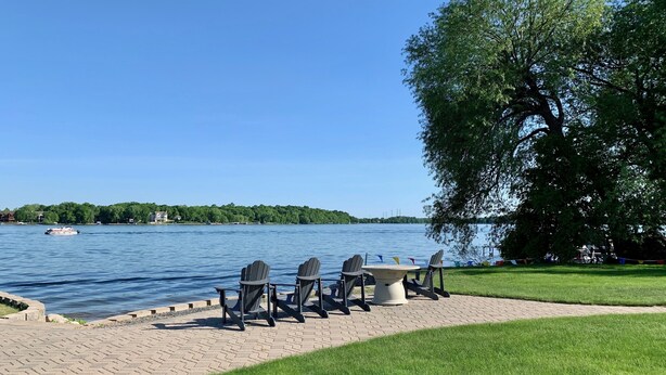 A lakeside patio with a table and chairs, a boat on the water, and a tree in the background. - 6BR Alexandria Lakefront, FREE Pontoon, Designed for Groups (Alexandria)