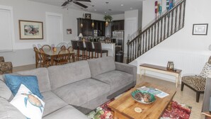 A living room with a gray sofa, wooden dining table, and chairs. A staircase with a wooden railing leads to an upper floor. - Spacious Home. Short Walk to Beach, Beach Shuttle & Shunk Gulley. Pet Friendly! (Santa Rosa Beach)