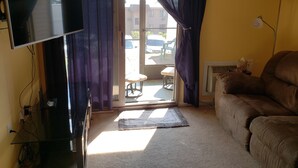 Un salon avec un téléviseur, un canapé, une table basse et une vue sur l’extérieur, visible à travers une porte vitrée.