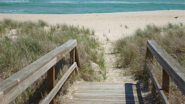 Een houten vlonder die naar een zandstrand leidt, met de oceaan op de achtergrond.