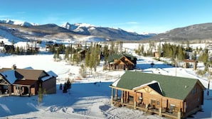 Uma cabana de tronco com varanda, rodeada por terreno e árvores cobertas de neve, com montanhas ao fundo.