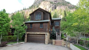 A two-story wooden house with a garage, a stone foundation, and a mountain backdrop.