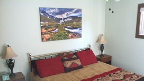 A bed with a patterned blanket, two bedside lamps, a wall-mounted painting of a landscape, and a nightstand with a lamp. - 10% Discount to Veterans, First Responders, and Seniors. Note status in request. (Estes Park)