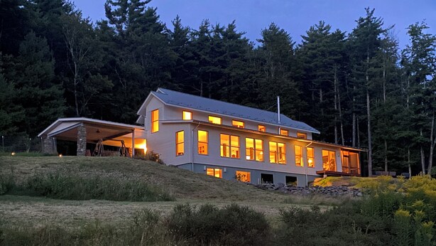 Una casa moderna de dos pisos con un amplio porche y numerosas ventanas, rodeada por un paisaje boscoso.
