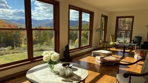 Un vaste salon doté de grandes fenêtres offrant une vue imprenable sur les montagnes et les couleurs automnales. Une table ronde, ornée d’un vase de fleurs et de bougies, est placée près des fenêtres.
