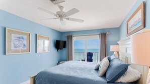 Un dormitorio con cama, ventilador de techo, televisión, ventana con vista al océano y un cuadro en la pared.