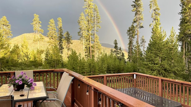 A wooden deck with a table and chairs, overlooking a forested hillside and a rainbow. - Excellent reviews. Perfect for ALL ages-families, kids, dogs allowed. Hot tub! (Grand Lake)