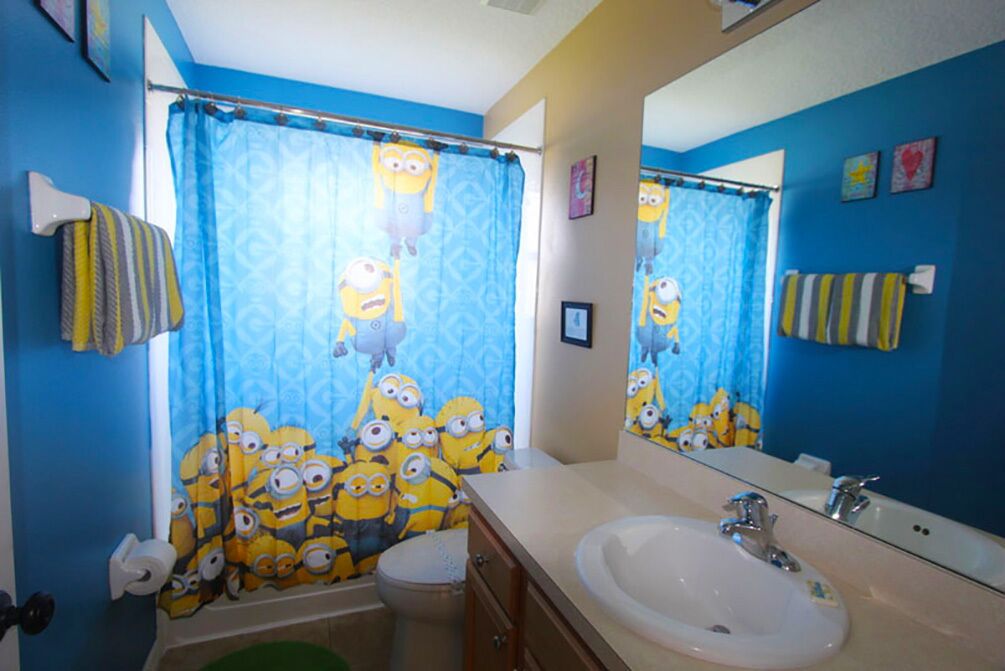 Un baño con cortina de ducha temática de Minions en azul y amarillo, un lavabo blanco y un espejo que refleja la decoración del baño.