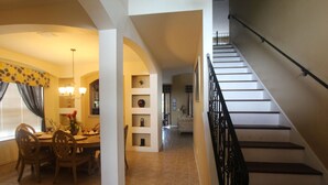 Un espace repas avec une table et des chaises, un escalier doté d’une rampe noire, et un couloir menant à un salon.