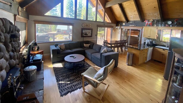 A spacious living area with a high ceiling, wooden beams, a large window overlooking a lake, a cozy seating area with a sofa and armchairs, and a modern kitchen with wooden cabinets and a stainless steel refrigerator. - Fun and Relaxing in Tahoe City! (Tahoe City)