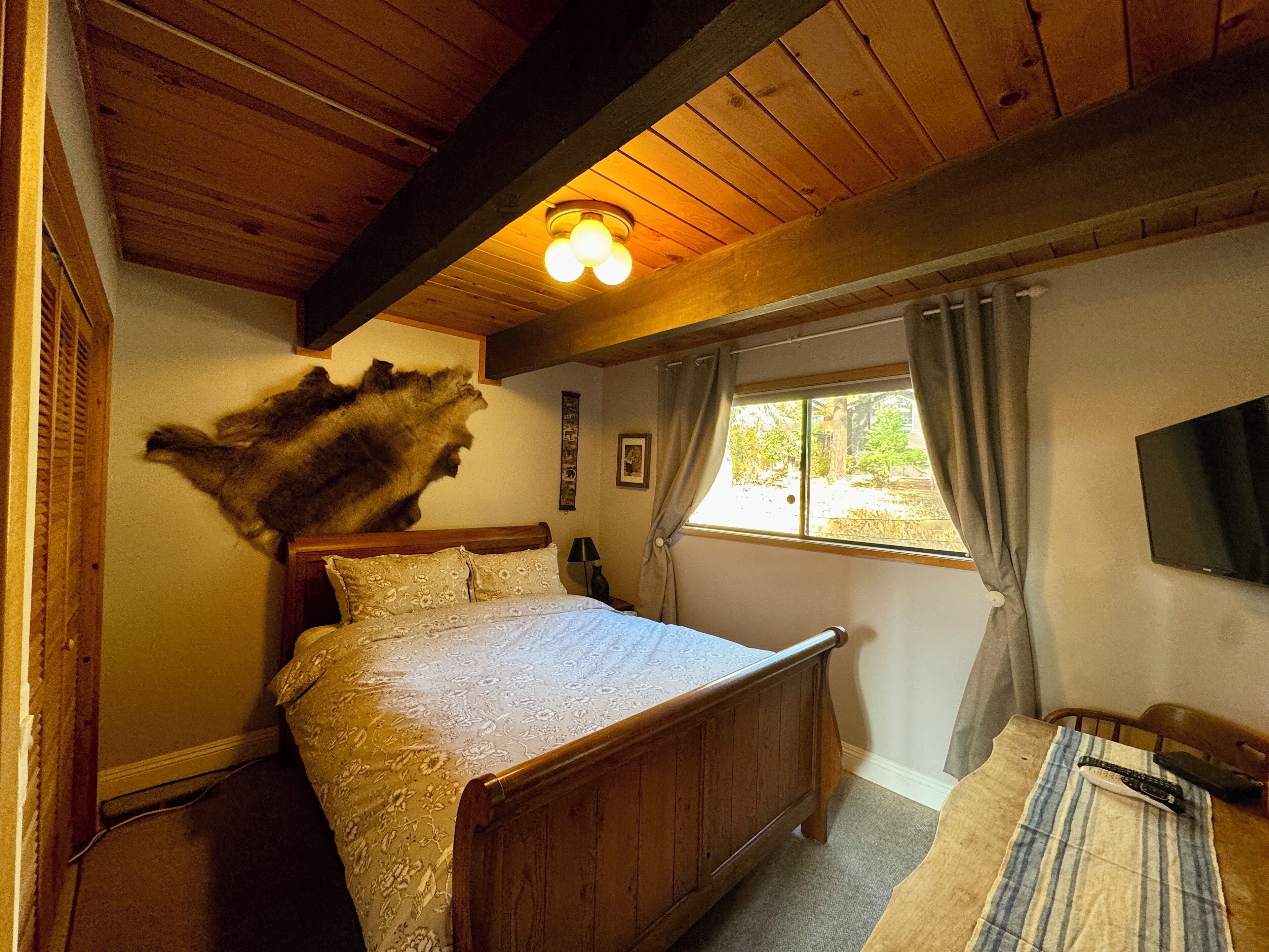 A wooden bedroom with a bed, a mounted television, and a window with curtains.