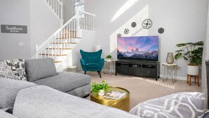 En moderne stue med en grå sofa, en turkis lænestol og et fladskærms-tv. En trappe med hvide gelændere ses i baggrunden.