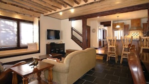 Una sala de estar con sofá, mesa de comedor de madera y sillas, televisión y una cocina con armarios de madera y una escalera.