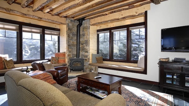 Una sala de estar acogedora con chimenea de piedra, vigas de madera, ventanas amplias y un televisor.