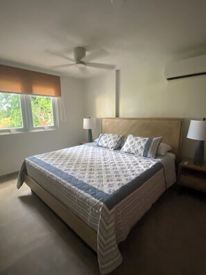 Ein Schlafzimmer mit einem Bett, einem Deckenventilator, einem Fenster mit Jalousien und einem Nachttisch mit Lampe.