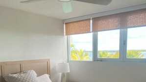 Ein Schlafzimmer mit einem Bett, einem Deckenventilator, einem Nachttisch mit Lampe und einem Fenster mit Blick auf Palmen.