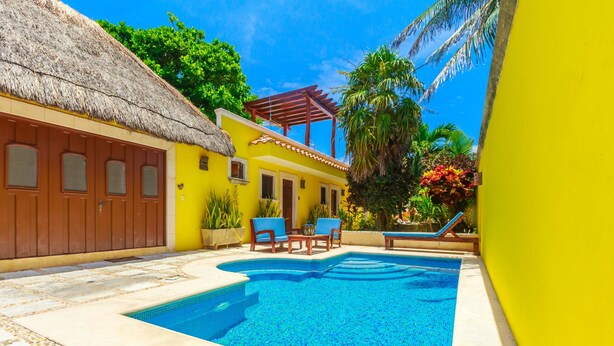 A swimming pool with a thatched roof structure, a yellow building, and a blue-tiled pool area. - One Bedroom Suite, Serene, Pool, Gardens, Rooftop Terrace with Views (Isla Mujeres)