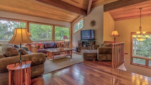 A spacious living room with wooden floors, large windows, a television, and a fireplace. - AC, Luxurious, vaulted ceilings, great home for couples, dogs allowed (Sunriver)