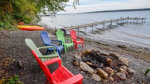 Ein Strand mit bunten Adirondack-Stühlen, einer Feuerstelle und einem Holzsteg, der sich über ruhiges Wasser erstreckt.
