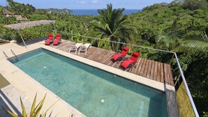 Una piscina con sillas reclinables rojas y un muelle de madera, rodeada de vegetación exuberante y vistas al océano.