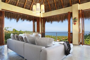 A modern living room with a sofa, a view of the ocean, and a thatched roof ceiling.