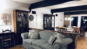 Een gezellige woonkamer met een geruit bankje, een houten eettafel en een keukenhoek met een bakstenen muur en moderne verlichting.
