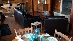 Interior acogedor de una cabaña con comedor que presenta una mesa redonda de madera dispuesta con copas y platos azules, rodeada de sofás de cuero y una mesa de comedor de madera.