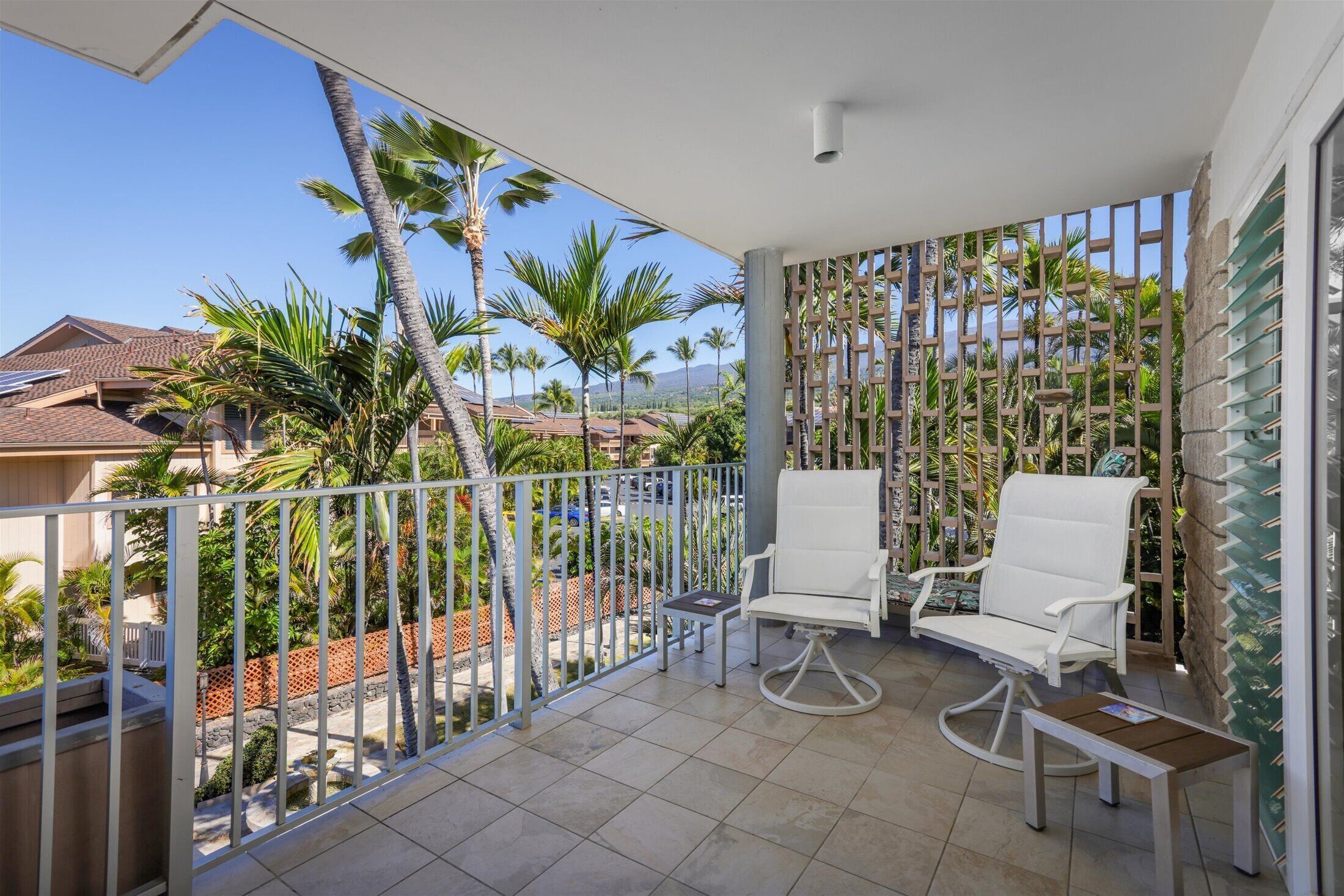 A balcony with two white chairs and a small table, overlooking a residential area with palm trees and houses.