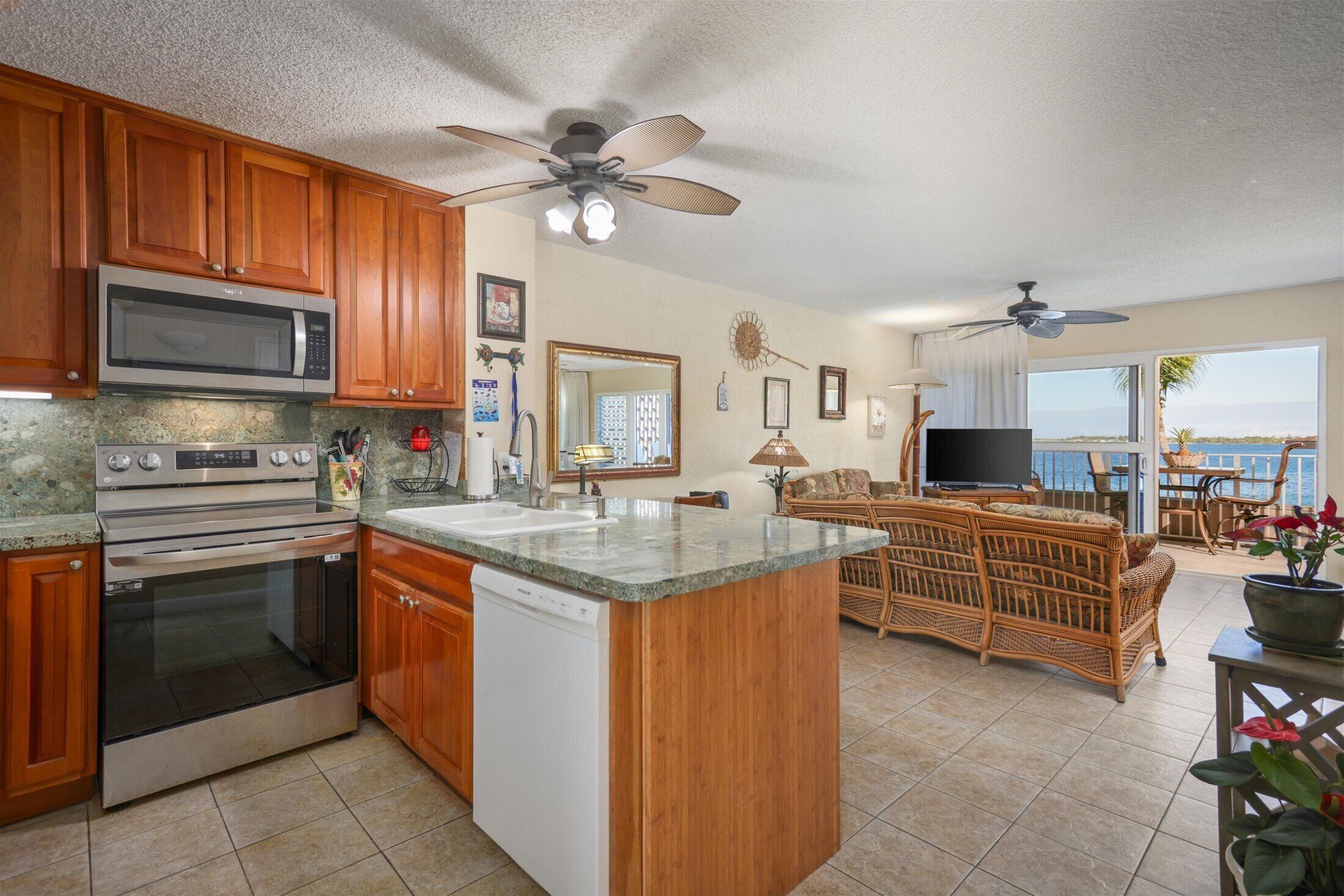 A kitchen with wooden cabinets, a microwave, an oven, and a dishwasher. There is a dining area with a wicker sofa and a TV.