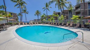 Una piscina con sillas de descanso y palmeras, rodeada por una cerca y edificios.