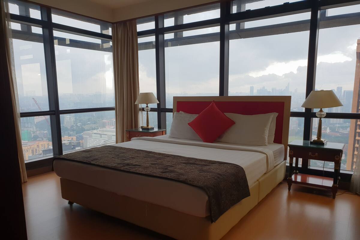 A large bed with a red headboard, two bedside tables with lamps, and a view of a cityscape through large windows.