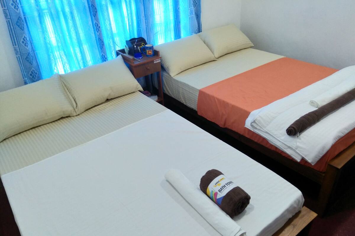 Dos camas individuales con sábanas blancas, una mesita de noche de madera con artículos de aseo, y una cortina azul al fondo.